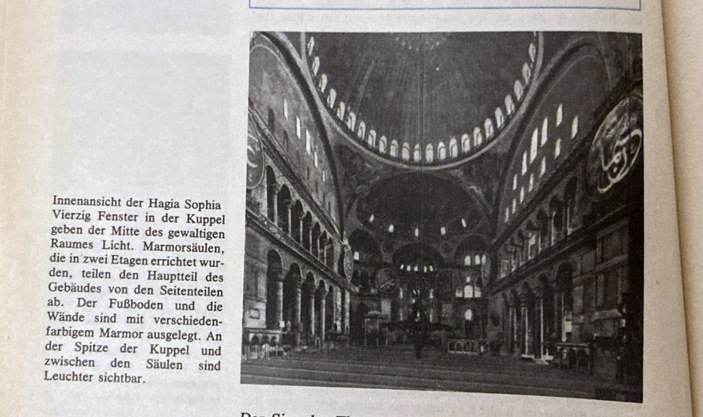 Innenansicht der Hagia Sophia mit Sicht auf die Kuppel, die von zahlreichen Fenstern erleuchtet wird. Marmorsäulen teilen den Raum in zwei Etagen. Leuchter hängen an der Spitze der Kuppel.